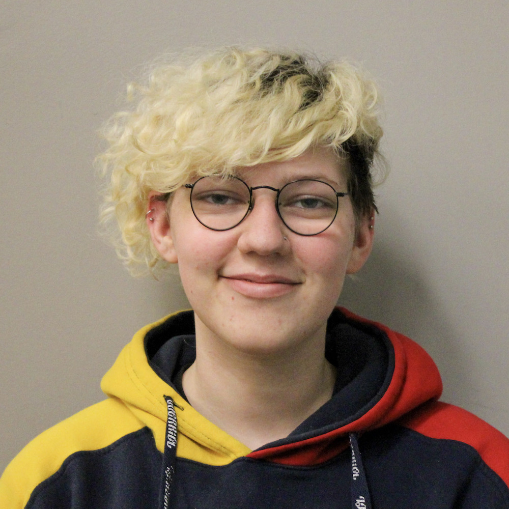 Daniel Braun has long bleach-blonde curly bangs and is wearing a yellow, black, and red hoodie and glasses.