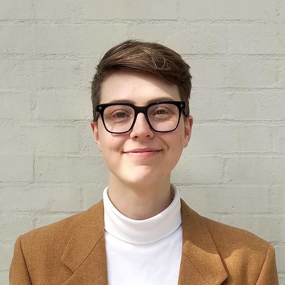 Orvis is a white non-binary person wearing glasses, a white turtleneck, and a cognac-coloured blazer. They stand in front of a white wall smiling at the camera.