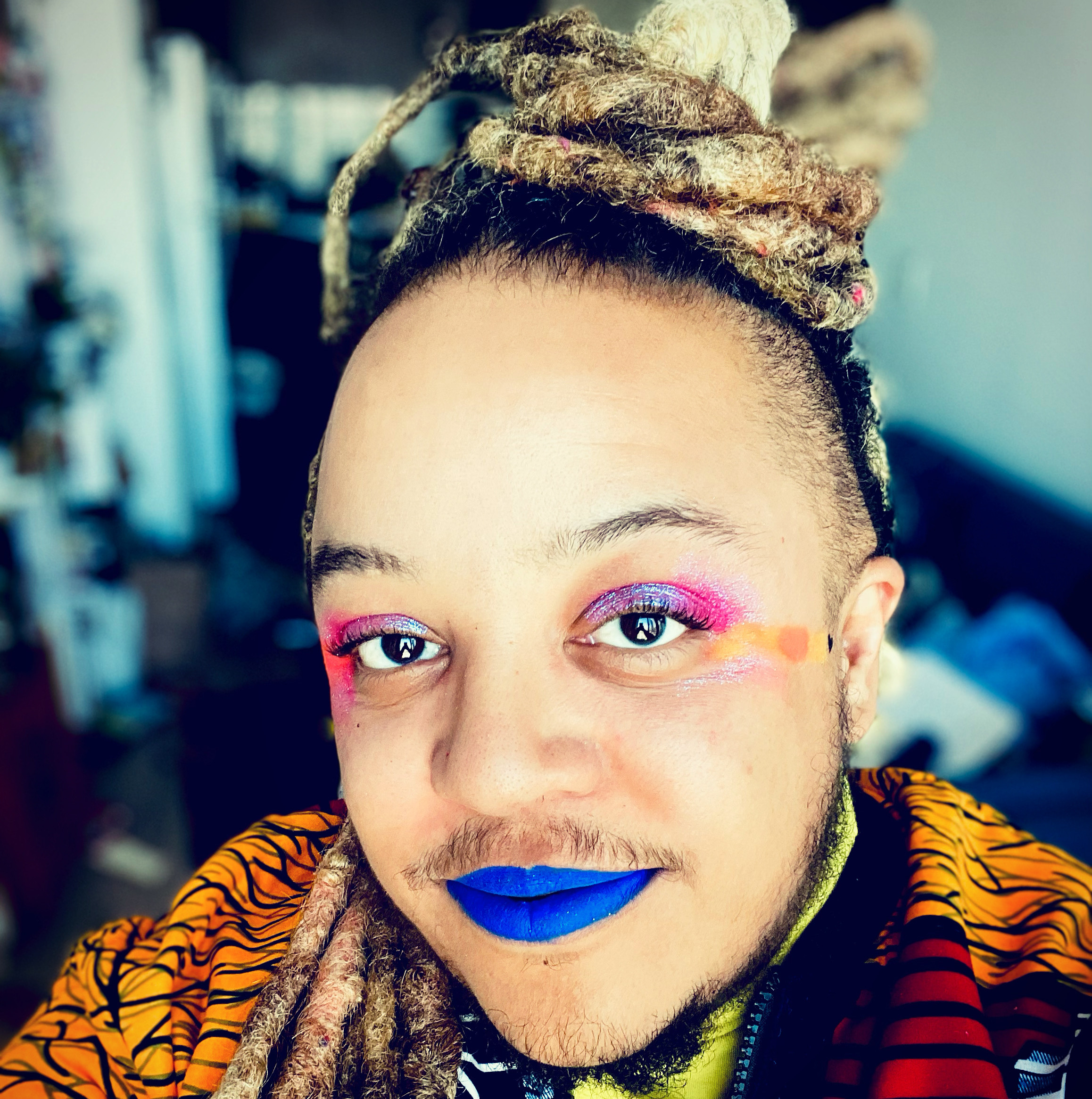 Syrus smiles up at the camera. He wears blue lipstick and pink, blue, and orange eyeshadow. His blonde dreadlocks are tied up and he wears a colourful jacket.